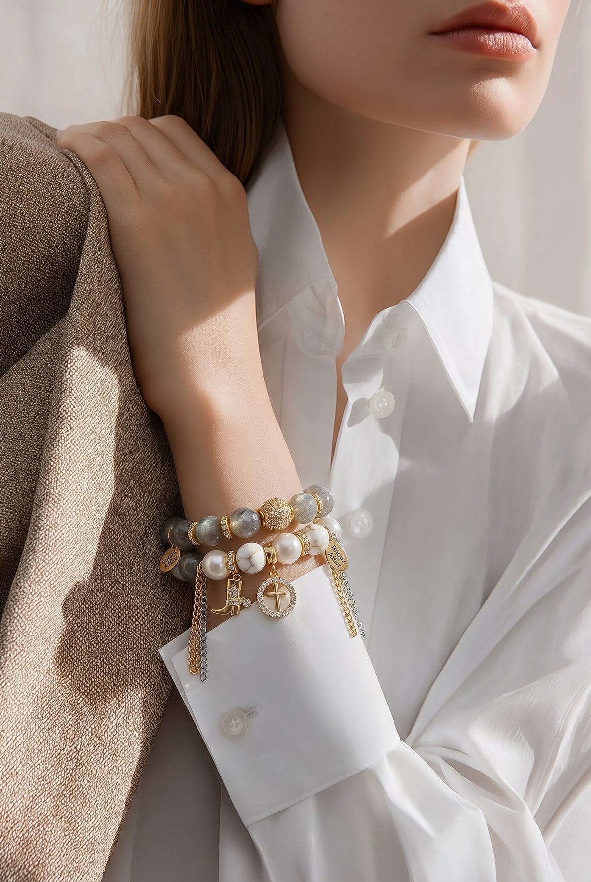 A woman in a white shirt with her hand on her shoulder, wearing two Bijoux Affair bracelets, grey labradorite and white pearl with gold chains, a cowboy boot charm and a crystal cross charm