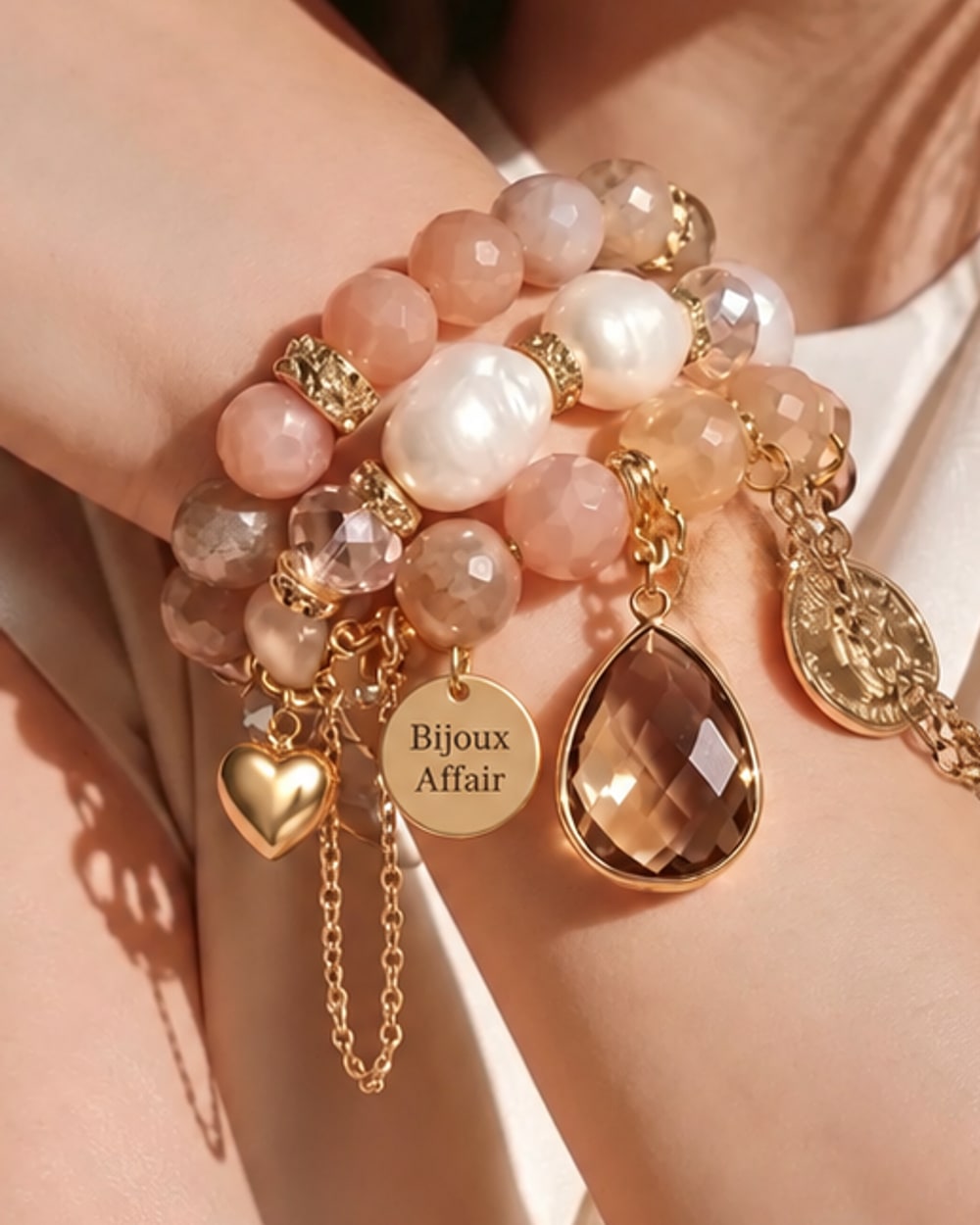 A wrist stacked with blush and peach gemstone bracelets, pearls, a smoky teardrop, a gold heart, a coin charm, dangling gold chains and the Bijoux Affair tag