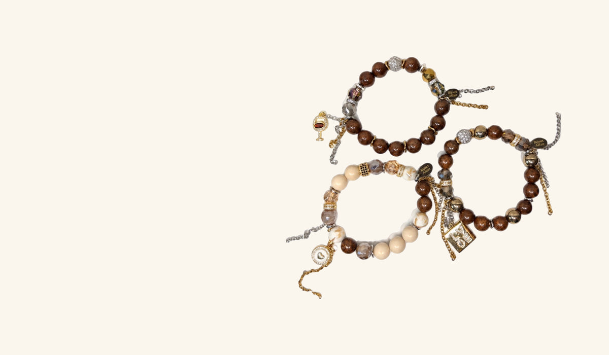 Three Bijoux Affair bracelets laid together on cream: brown wood and crystal beads with a wine glass charm, brown wood and gold hematite with a gold LOVE charm, and cream stone with brown accents and an enamel heart charm. Shown wide as the New Arrivals banner