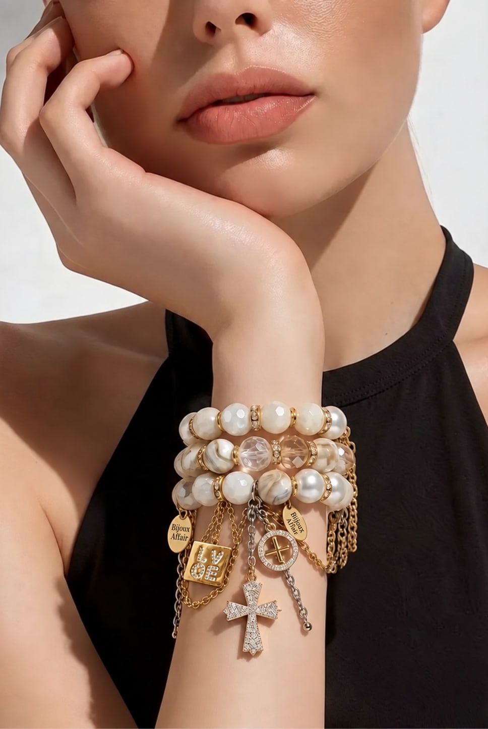 A woman in a black halter top resting her chin on her hand, wearing a BIJOUXstacks set of three white pearl and crystal bracelets with gold chains, Bijoux Affair tags, a LOVE charm and a crystal cross