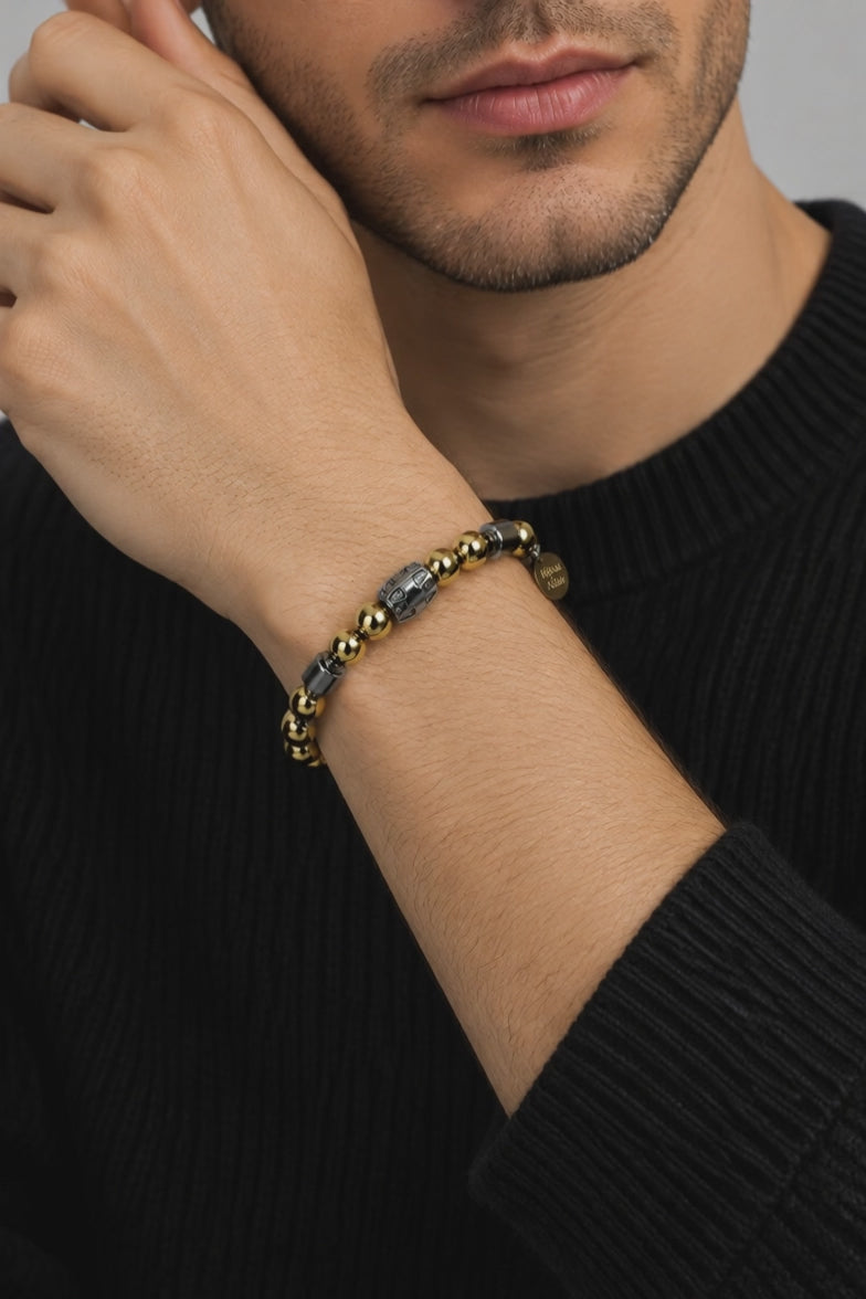 A man in a black knit sweater, hand raised to his face, wearing a gold and hematite bead bracelet with a round gold tag